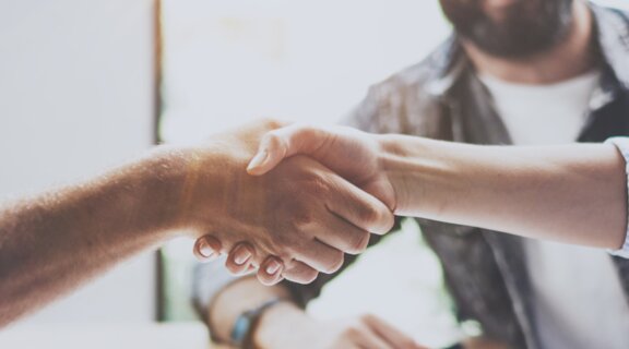 Drei Menschen sitzen am Tisch, zwei geben sich die Hände, alle lächeln.